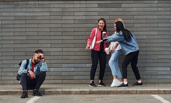 Three women standing and laughing at guy that sitting outdoors near building at daytime.