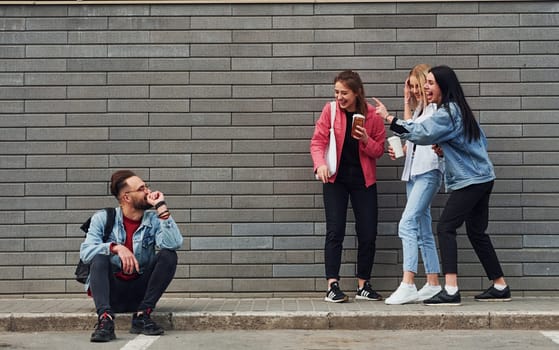 Three women standing and laughing at guy that sitting outdoors near building at daytime.