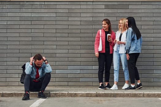 Three women standing and laughing at guy that sitting outdoors near building at daytime.