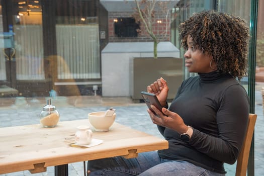 young african american woman sitting in a cafe with a cup of hot coffee, watching a social media feed in a smartphone, using technology, high quality photo, High quality photo