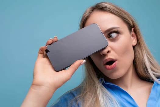 close-up of a shocked european blonde girl holding a smartphone screen with a mockup for an application near her face on a blue background.