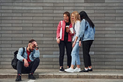 Three women standing and laughing at guy that sitting outdoors near building at daytime.