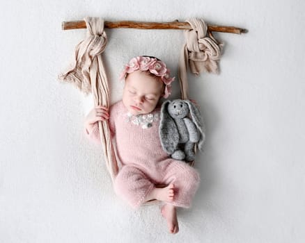 Newborn baby girl sleeping holding knitted bunny toy in swing decoration. Cute infant child kid napping studio portrait