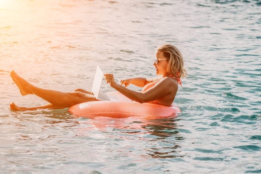 Woman works on laptop in sea. Freelancer, young blond woman in sunglases floating on an inflatable big pink donut with a laptop in the sea at sunset. Freelance, travel and holidays concept