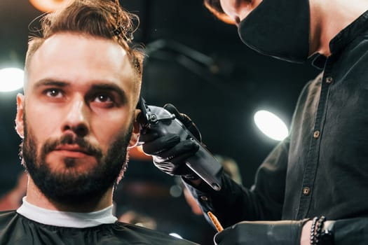 Front view of young bearded man that sitting and getting haircut in barber shop by guy in black protective mask.