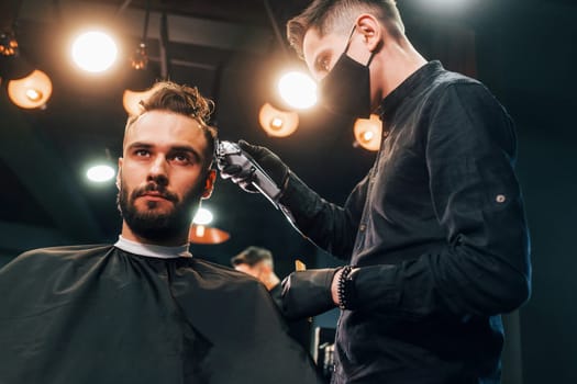 Front view of young bearded man that sitting and getting haircut in barber shop by guy in black protective mask.