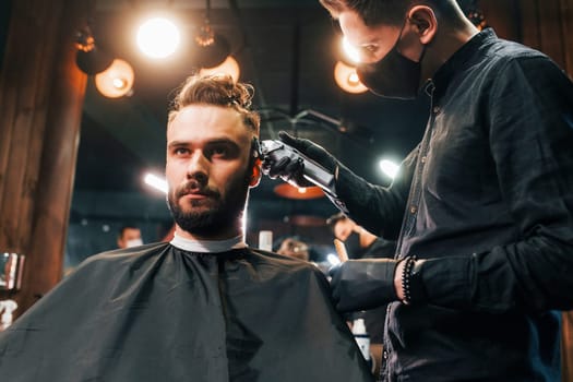 Front view of young bearded man that sitting and getting haircut in barber shop by guy in black protective mask.