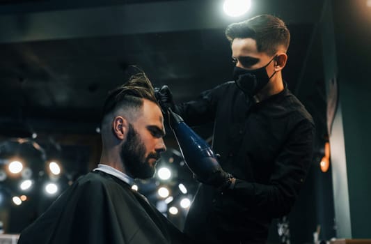Young bearded man sitting and getting haircut in barber shop by guy in black protective mask.