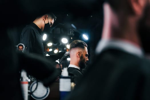 Young bearded man sitting and getting haircut in barber shop by guy in black protective mask.