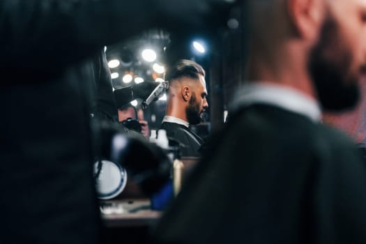 Young bearded man sitting and getting haircut in barber shop.