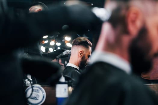 Young bearded man sitting and getting haircut in barber shop.