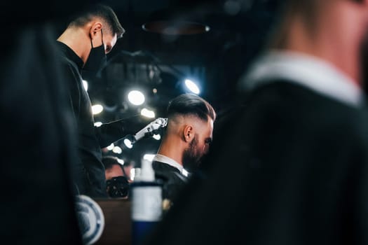 Young bearded man sitting and getting haircut in barber shop.
