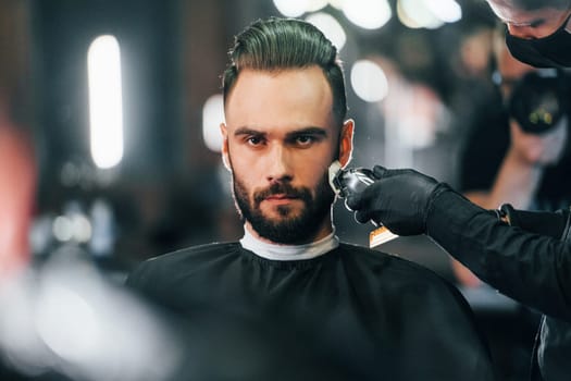 Young man with stylish hairstyle sitting and getting his beard shaved in barber shop.