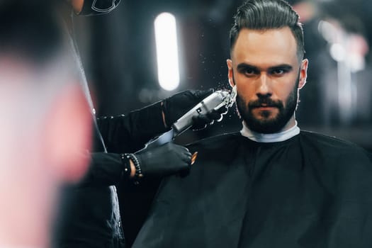 Young man with stylish hairstyle sitting and getting his beard shaved in barber shop.