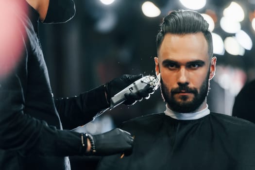 Young man with stylish hairstyle sitting and getting his beard shaved in barber shop.