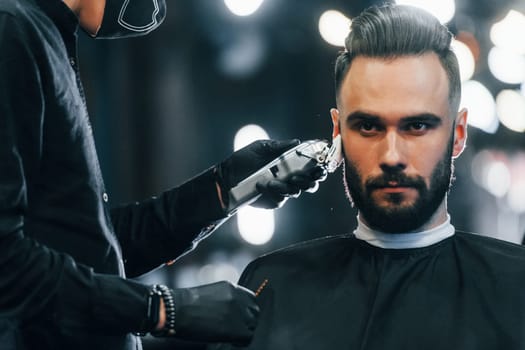 Young man with stylish hairstyle sitting and getting his beard shaved in barber shop.