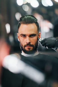 Young man with stylish hairstyle sitting and getting his beard shaved in barber shop.