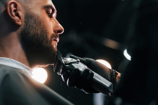 Young man with stylish hairstyle sitting and getting his beard shaved in barber shop.