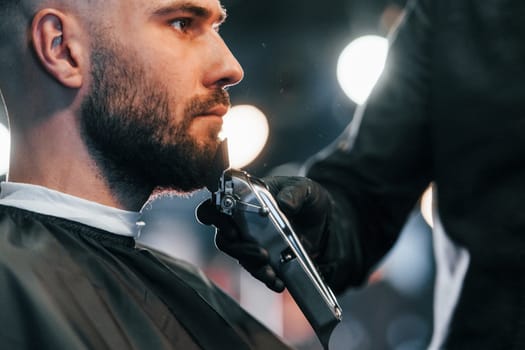 Young man with stylish hairstyle sitting and getting his beard shaved in barber shop.