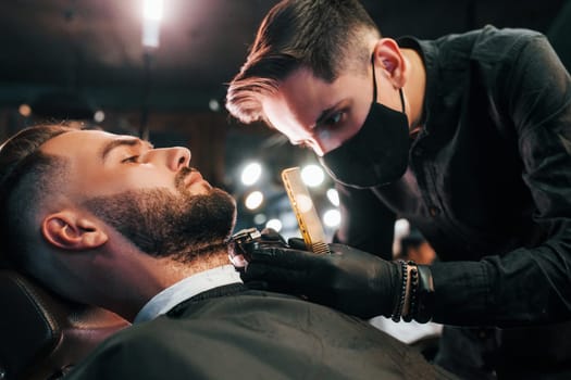 Young man with stylish hairstyle sitting and getting his beard shaved by guy in black protective mask in barber shop.