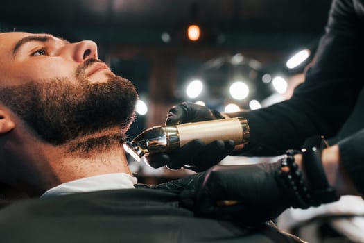 Young man with stylish hairstyle sitting and getting his beard shaved in barber shop.