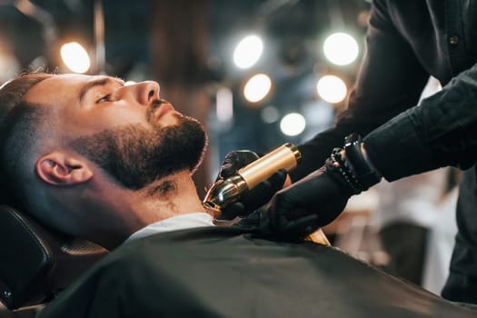 Young man with stylish hairstyle sitting and getting his beard shaved in barber shop.