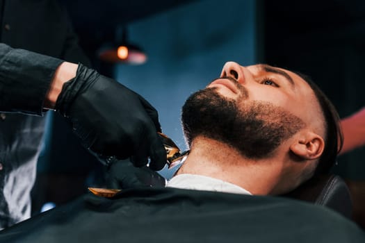 Young man with stylish hairstyle sitting and getting his beard shaved in barber shop.