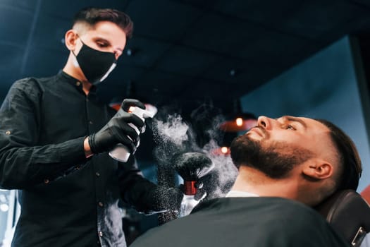 Young man with stylish hairstyle sitting and getting his beard shaved by guy in black protective mask in barber shop.