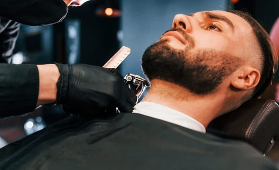 Young man with stylish hairstyle sitting and getting his beard shaved in barber shop.