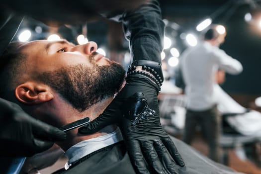 Young man with stylish hairstyle sitting and getting his beard shaved in barber shop.