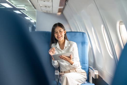 Young asian businesswoman in formal clothes working using tablet with smart pen while sitting in airplane cabin near window traveling to another place.