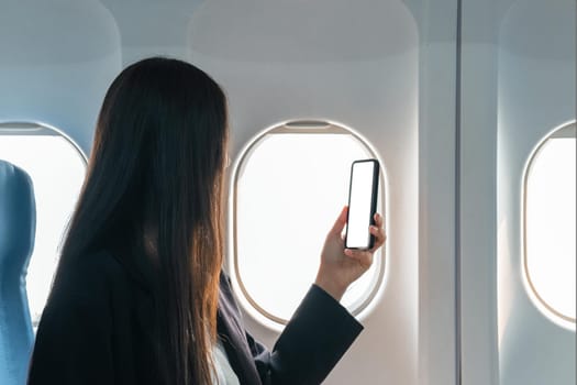 A female traveler passenger sits at the window seat in economy class, using his smartphone, holding a mobile phone white screen mockup. close-up image.