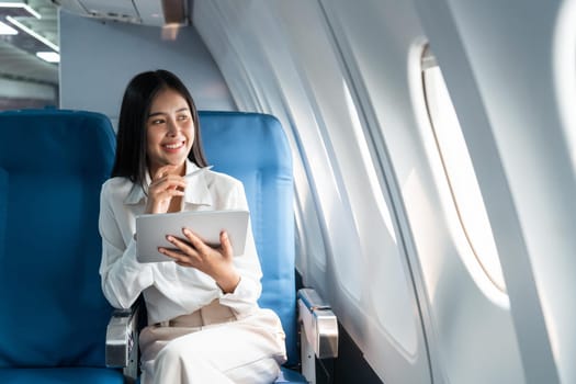 Young asian businesswoman in formal clothes working using tablet with smart pen while sitting in airplane cabin near window traveling to another place.