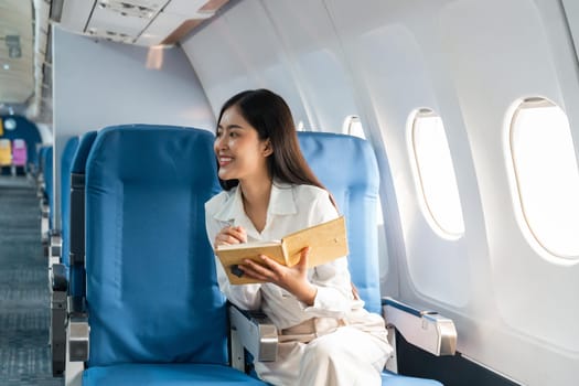 Smiling Asian woman taking notes of information and sitting in airplane, Business traveling and technology concept.