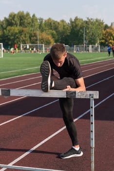 Young sporty man athlete runner in sportswear stretching before running hurdles on stadium