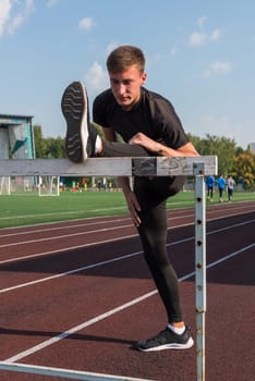 Young sporty man athlete runner in sportswear stretching before running hurdles on stadium