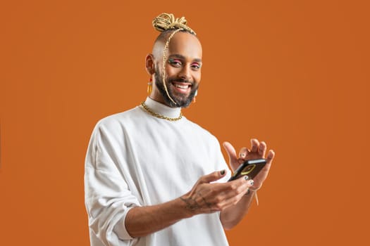 Confident black gay man with bright makeup use smartphone standing isolated on orange background, dressed white. Exudes sense of pride and individuality. Diversity power of personal style. Side view