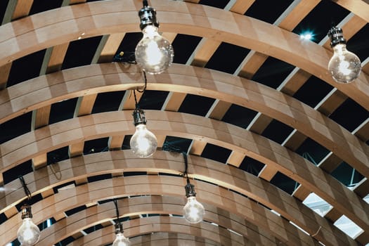 Many White Light Bulbs are hung on the Ceiling of the Wooden Curved Pergola Structure. Light Bulbs in Wooden Facade Architecture. Space for text, Selective focus.