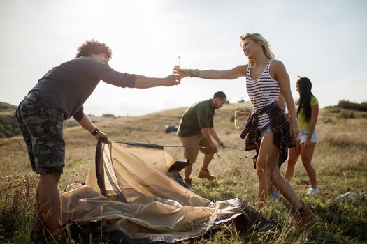 Group of a cheerful young friends putting up their tent on a suitable place in a meadow.