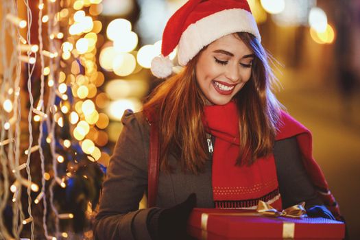 Shoot of a smiling young woman with red present box is having fun in the city street at Christmas time.