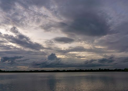 The clouds cover the sky, obscuring the light from the sun at sunset. The skyline divides the pond and the sky is covered with trees.