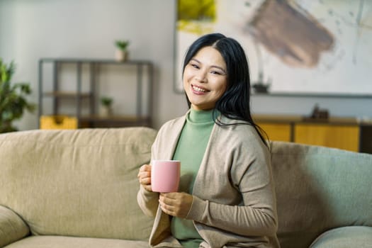 mature Asian woman enjoys a hot cup of tea while relaxing on her cozy living room sofa. High quality photo