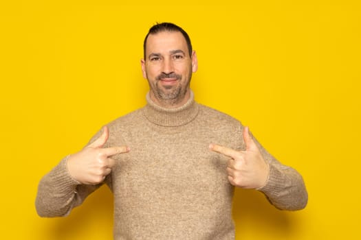 The portrait of a bearded handsome man points to himself puzzled at being chosen. Amazed and attractive hispanic man over yellow background, he has a confused expression