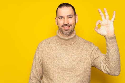 Bearded hispanic man in his 40s wearing a beige turtleneck making the ok gesture while smiling at the camera, isolated over yellow background