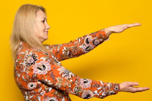 Caucasian blonde woman holding something with both hands in profile isolated on yellow background. Copy space between hands for advertising, text or objects.