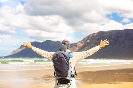 Young father rising hands to the sky while enjoying pure nature carrying his infant baby boy son in backpack on windy sandy beach of Famara, Lanzarote island, Spain. Family travel concept