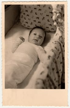 GERMANY - 1954: Vintage photo shows cute small baby in a cot - crib, baby bed. Retro black and white photography. Circa 1950s.