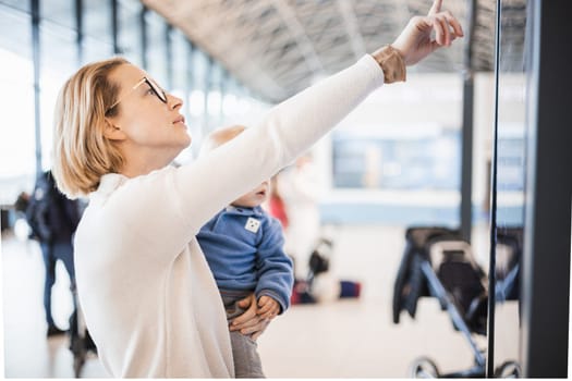 Mother traveling with child, holding his infant baby boy at airport terminal, checking flight schedule, waiting to board a plane. Travel with kids concept