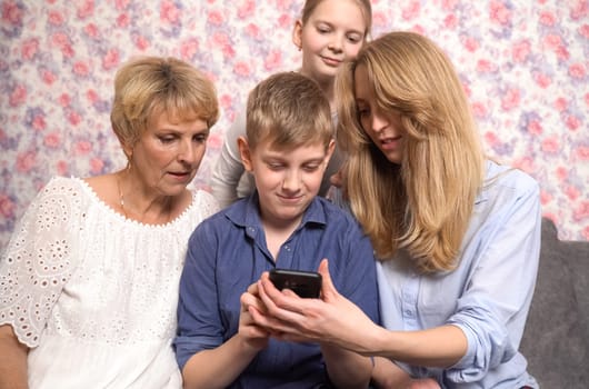 Happy grandchildren, grandmother and mother have fun using smartphone, sitting on sofa.