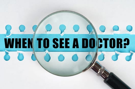 Between two sheets from a notebook on a blue stripe with the inscription - When to see a doctor, there is a magnifying glass. Medical concept.
